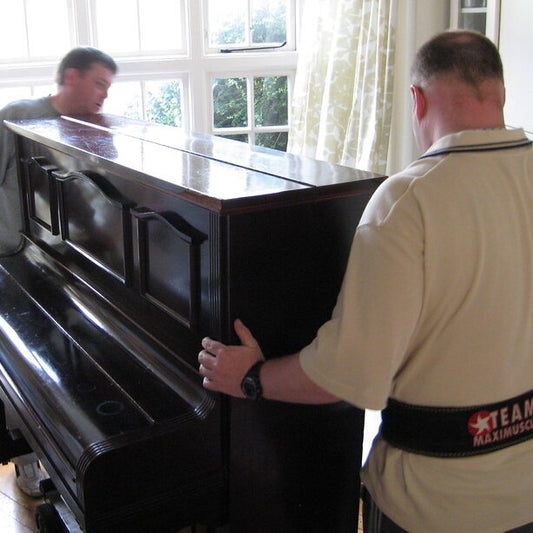 Two professional movers from Nomad Moving Company carefully maneuvering an upright piano in a dining room, showcasing their expertise in piano and safe moving services in Lexington and Louisville, KY.